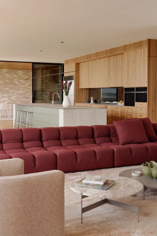 A warm, open-plan living and kitchen interior with a large deep red modular sectional sofa in the foreground. Behind it is a contemporary timber-lined kitchen with full-height wood cabinetry, integrated dark appliances, a pale stone island benchtop, and a tiled feature wall. A marble-look oval coffee table, neutral upholstered armchair, and soft beige rug complete the refined, richly coloured space.