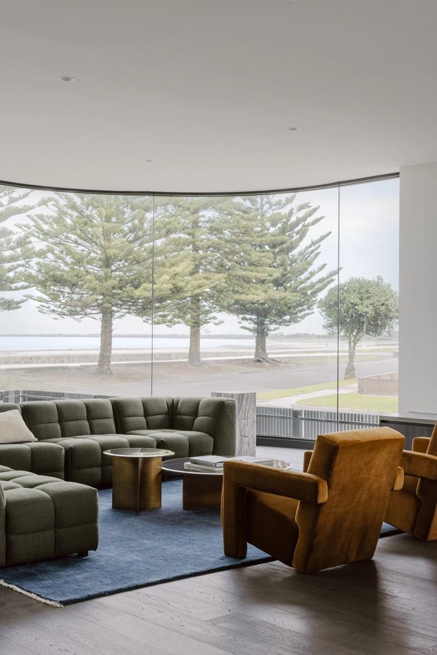 A refined living room with a large curved wall of floor-to-ceiling glazing overlooking a tree-lined waterfront or coastal promenade. The room features a deep olive-green modular sectional, cognac-brown lounge chairs, metallic cylindrical coffee tables, a dark blue area rug, and pale timber flooring. The interior is softly lit and spacious, with a calm, sophisticated atmosphere.