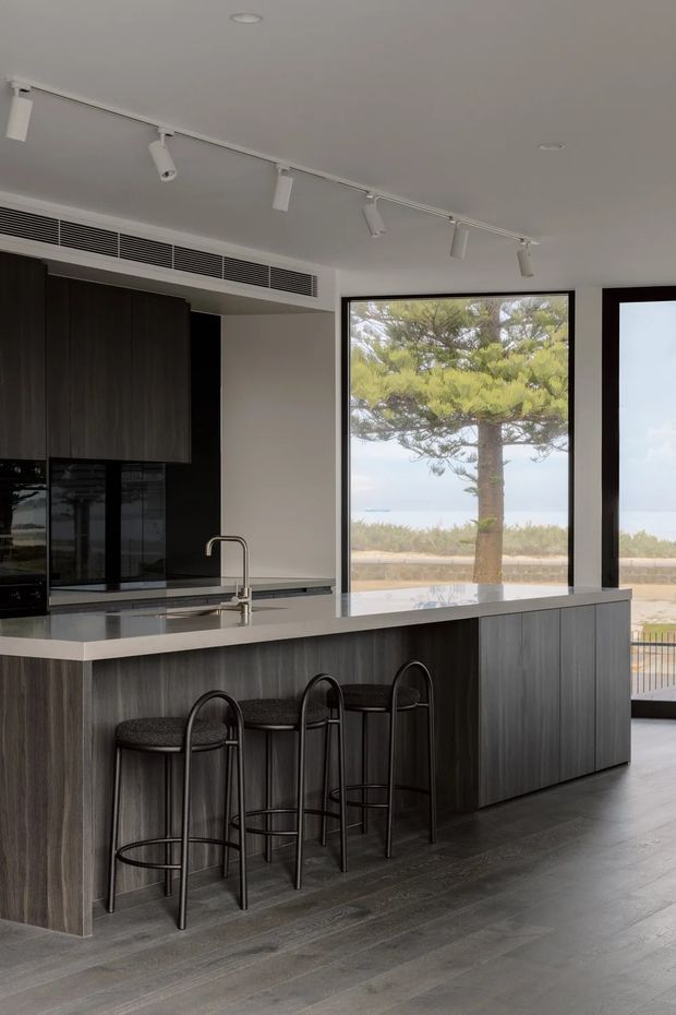 Contemporary kitchen interior with a large dark wood-grain island, dark cabinetry, a pale stone benchtop, integrated appliances, and three black bar stools. Track lighting and a linear air-conditioning grille run across the ceiling. Full-height glazing provides views toward trees, water, and a balcony or terrace beyond.