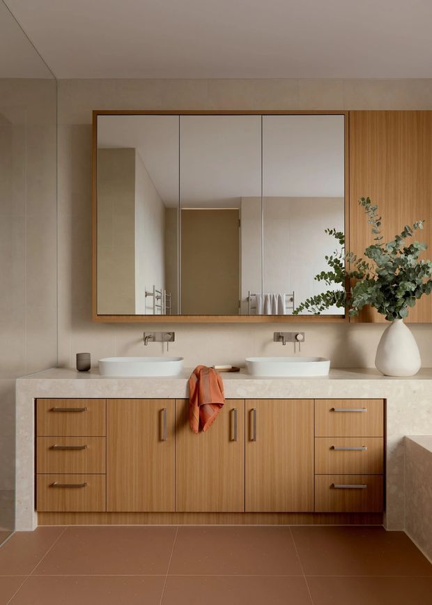 A refined contemporary double-vanity bathroom with a warm timber floating vanity, pale stone countertop, two white vessel basins, wall-mounted brushed metal tapware, and a large frameless mirror. A vertical timber feature panel and leafy vase arrangement add warmth, while large brown floor tiles and neutral walls create a calm, earthy palette.