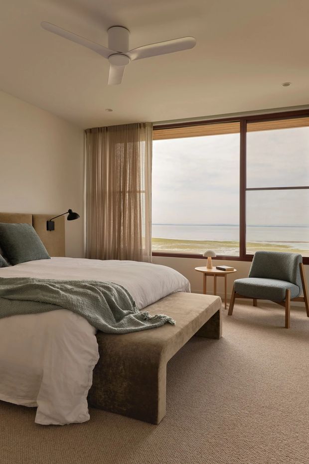 A calm, warm-toned bedroom with a large bed dressed in white linen and a textured grey-green throw, a substantial upholstered bench at the foot, and a matching upholstered timber-framed armchair. Full-height curtains frame wide timber-framed glazing overlooking a beach and ocean. A ceiling fan, wall-mounted reading light, and small round timber side table complete the understated coastal interior.