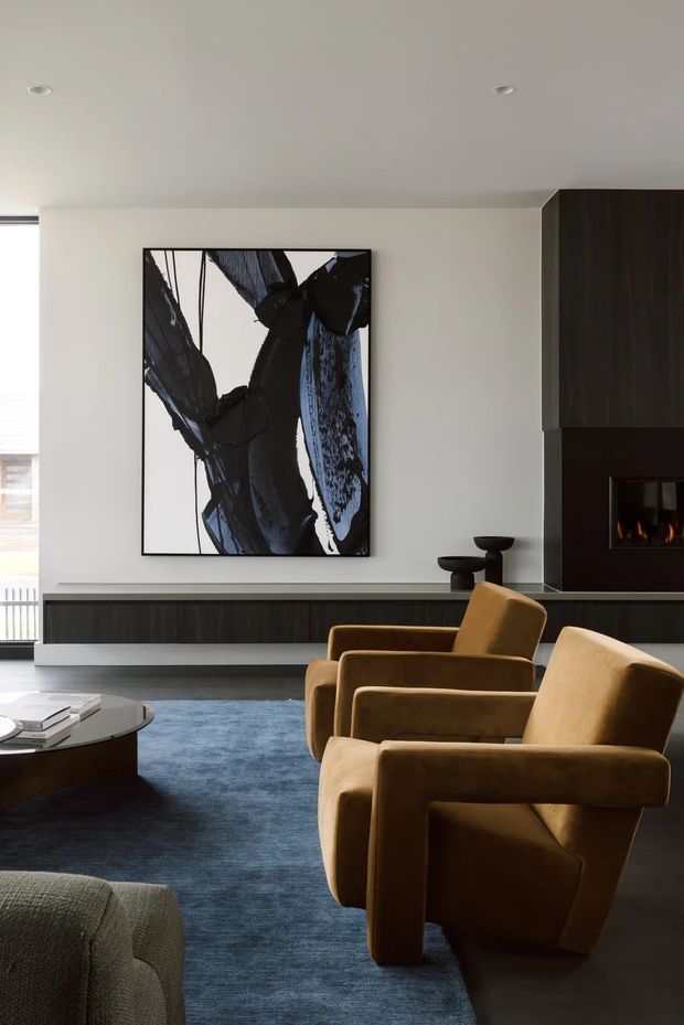 A refined contemporary living room with two mustard-brown upholstered armchairs facing a low dark coffee table, a deep blue rug, and a dark built-in media wall with a linear fireplace. The room has pale walls, a smooth white ceiling with recessed downlights, dark timber-toned cabinetry, and a large monochrome abstract artwork. Daylight enters from glazing on the left, creating a calm, sophisticated interior.