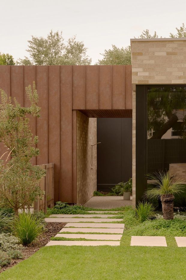 A landscaped residential entry courtyard viewed from outdoors, with a narrow stepping-stone path through lawn and low planting leading to a recessed doorway. The facade combines rust-coloured vertical metal panels, warm tan masonry, and a dark-framed glazed opening. The planting is restrained and contemporary, with ornamental grasses, shrubs, and a small sculptural palm-like plant. The composition has a warm, earthy, minimalist contemporary character.