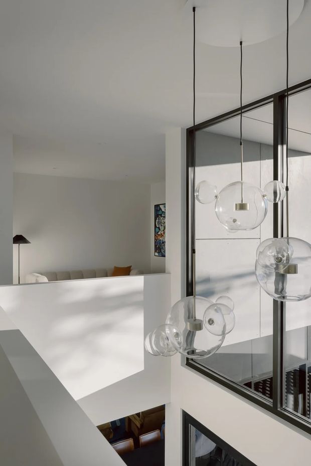 A bright, minimalist interior viewed across an open void, featuring a sculptural cluster of suspended glass globe pendant lights with dark cords and brass-toned fittings. Large dark-framed internal glazing overlooks a lower-level dining or living area, while a white sitting area and abstract artwork are visible beyond. Strong natural light creates geometric shadows across the white surfaces.