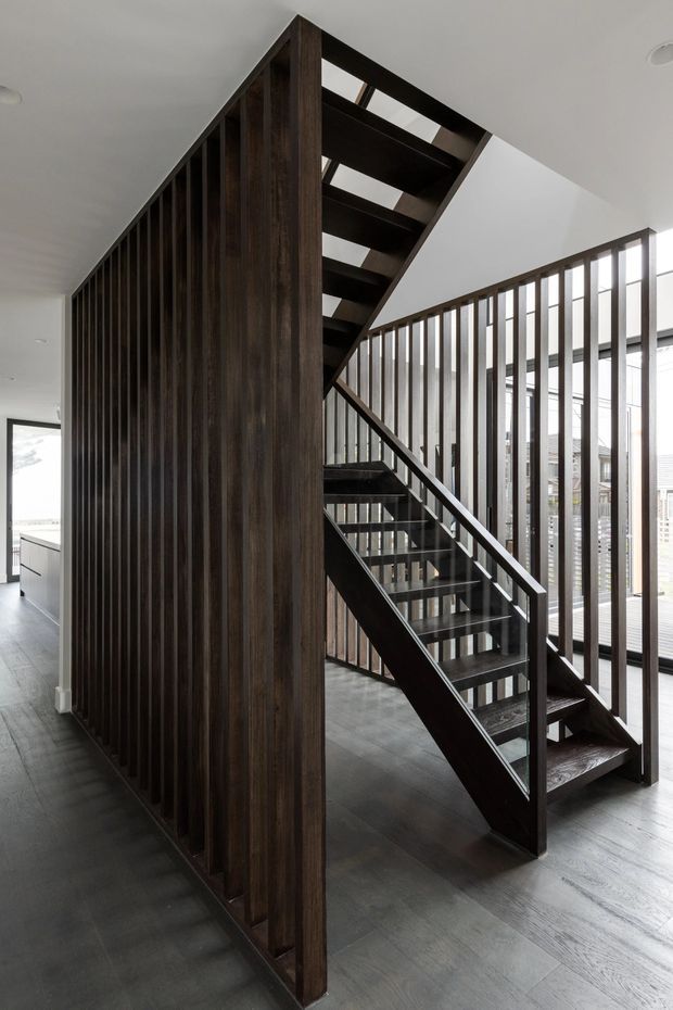 A contemporary interior stairway with dark timber open-riser stairs, matching vertical timber slat screens, and slim balustrades. The stair rises through a bright, double-height space with large windows, white ceilings, and dark flooring, creating a strong geometric contrast between the timber structure and natural daylight.