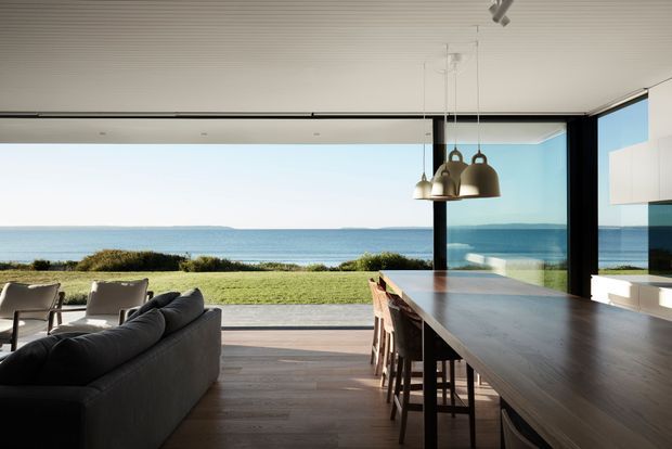 Open-plan contemporary living and dining space with a long timber kitchen island, bar stools, dark sectional sofa, and sculptural pendant lights. Full-height glazing opens directly to a lawn and expansive ocean view, creating a bright, minimalist coastal setting. The interior features dark-framed glazing, timber flooring, and a linear white ceiling.
