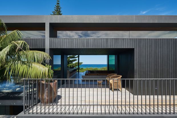 Exterior view of a contemporary flat-roofed residence with dark vertical cladding, exposed concrete beams, broad glazing, and a raised terrace with slender metal balustrade. A palm tree and ocean horizon are visible beyond the house, giving the setting a coastal character.