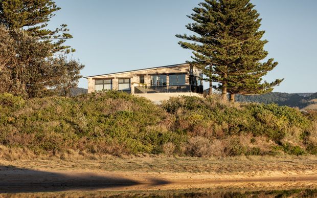 A low, contemporary single-storey house set on a vegetated coastal or rural slope. The building has broad glazing, a dark horizontal balcony or terrace edge, pale masonry or stone walls, and a shallow low-pitched roof. Native-looking trees and scrub surround the home, with distant hills visible beyond.