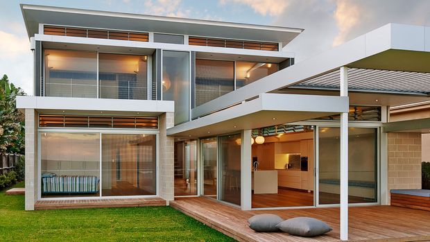 Exterior view of a modern multi-level residence with strong horizontal roof planes, extensive floor-to-ceiling glazing, covered outdoor entertaining deck, and an open-plan kitchen visible through the glass. The facade combines white rendered surfaces, light brickwork, timber screening, and warm interior lighting, with a small lawn in the foreground.