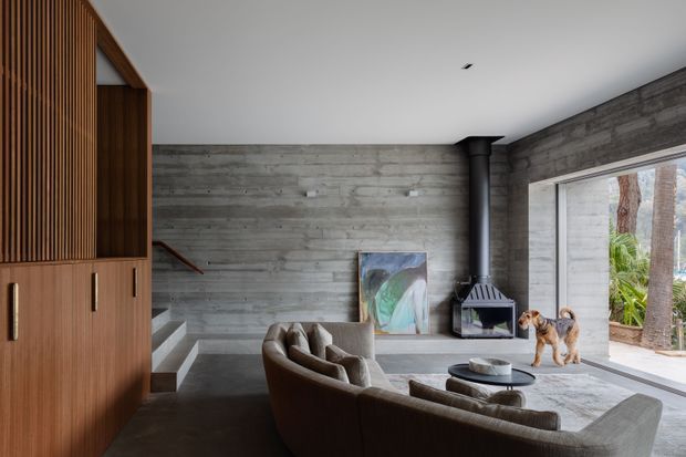 A contemporary living room with exposed horizontal concrete walls, a large open glazed doorway to an outdoor area, and a sculptural black wood-burning fireplace. Curved brown upholstered sofas surround a round coffee table on a pale rug. Vertical timber slat screening and matching built-in cabinetry form a feature wall on the left, while a stair descends behind the seating. The space has a restrained, warm-modern palette with strong textural contrast between concrete, timber, and upholstery.