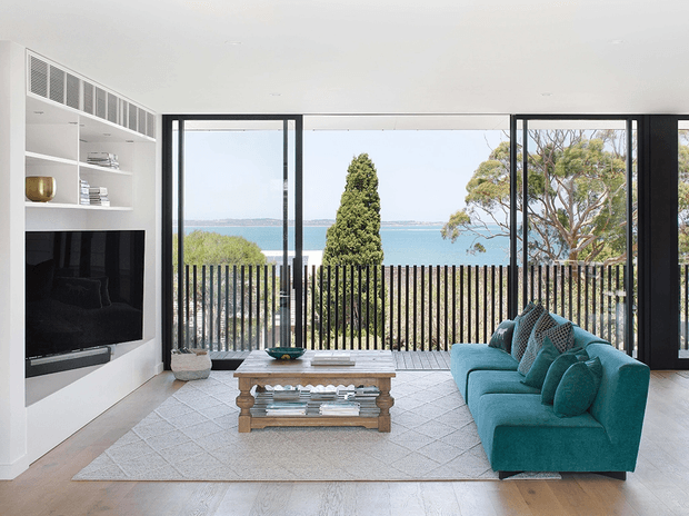 Bright contemporary living room with a large teal sofa, built-in white media cabinetry and shelving, a timber coffee table, and pale textured flooring. Full-height black-framed glazing opens to a balcony or terrace with a vertical slat balustrade and views toward blue water and coastal vegetation.