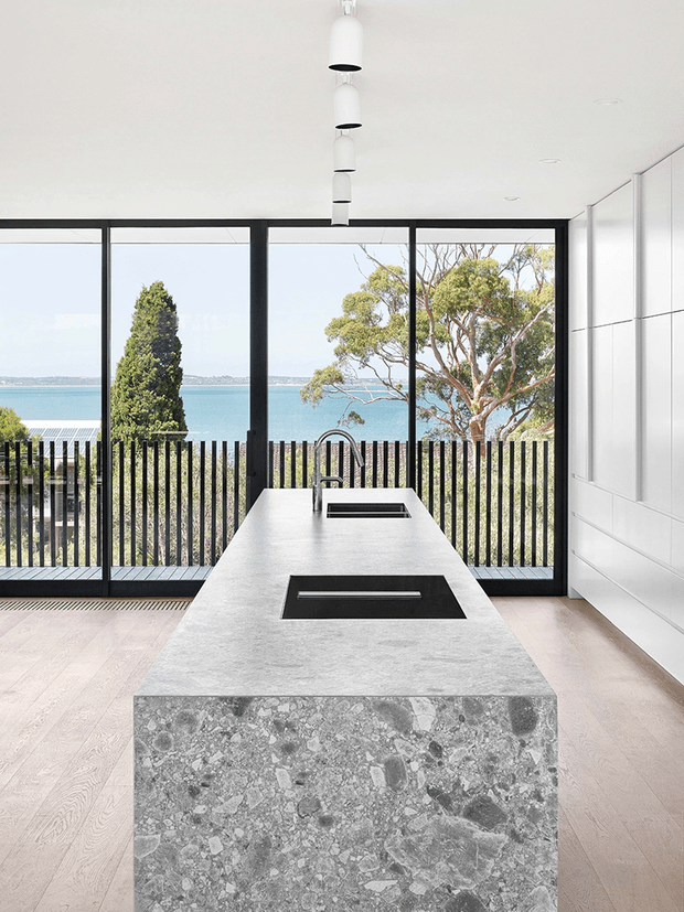 Contemporary kitchen interior centered on a large grey terrazzo island with an integrated sink and black cooktop. Full-height black-framed glazing opens to a balcony or terrace with a vertical metal balustrade and a visible coastal water outlook. White flat-panel cabinetry lines the right wall, while a row of pendant lights hangs above the island.