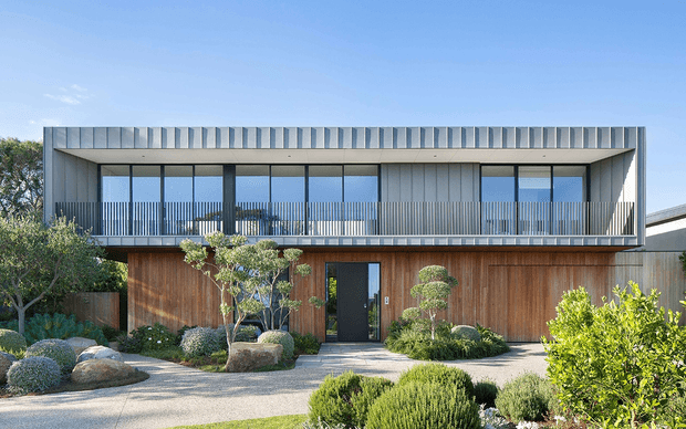 Contemporary two-storey residence with a highly rectilinear facade, upper-level full-height glazing, a deep covered balcony with slender metal balustrades, and a warm timber-clad lower level. The home is set within a landscaped garden featuring sculpted shrubs, rocks, and small ornamental trees.
