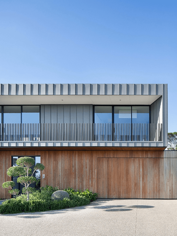 A contemporary two-storey residential facade with a warm vertically textured timber-clad lower level and a crisp upper level featuring extensive glazing, dark slender frames, and a screened balcony. The roofline has a repeating folded metal parapet detail, while sculpted planting and a pale paved driveway soften the frontage.