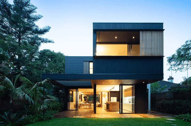 A contemporary multi-level residential house set within lush landscaped planting, photographed at dusk. The dark-clad rectilinear building features a projecting upper storey, broad glazed openings, a sheltered ground-floor terrace, warm timber soffits, and an illuminated open-plan interior. The composition highlights a strong indoor–outdoor connection and a flat-roofed, modern form.