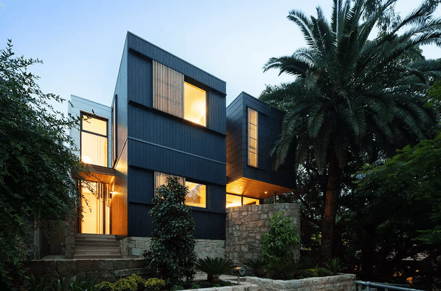 Exterior view of a contemporary multi-level residence composed of dark vertically clad geometric volumes, with warm illuminated glazing, projecting upper sections, and a rugged stone base and retaining wall. Dense tropical landscaping, including a prominent palm, frames the building.
