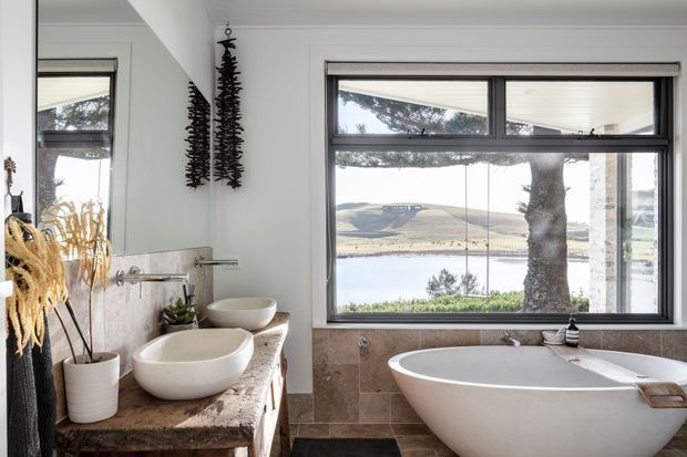 A bright, refined bathroom with a freestanding white bathtub positioned in front of a large black-framed window overlooking a lake and rolling hills. A stone-topped vanity with two vessel basins, wall-mounted taps, a large mirror, potted plants, and textured decorative elements completes the room. The palette is predominantly white, beige stone, dark grey, and black accents.