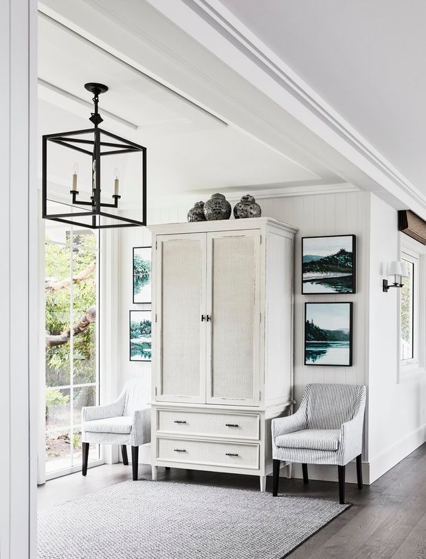 Bright, refined coastal-style interior featuring a white paneled room with decorative ceiling mouldings, a large built-in-looking freestanding armoire, two striped upholstered armchairs, framed landscape artwork, a geometric black lantern pendant, and pale textured rug over dark timber flooring. Tall windows and glazed doors bring in abundant natural light.