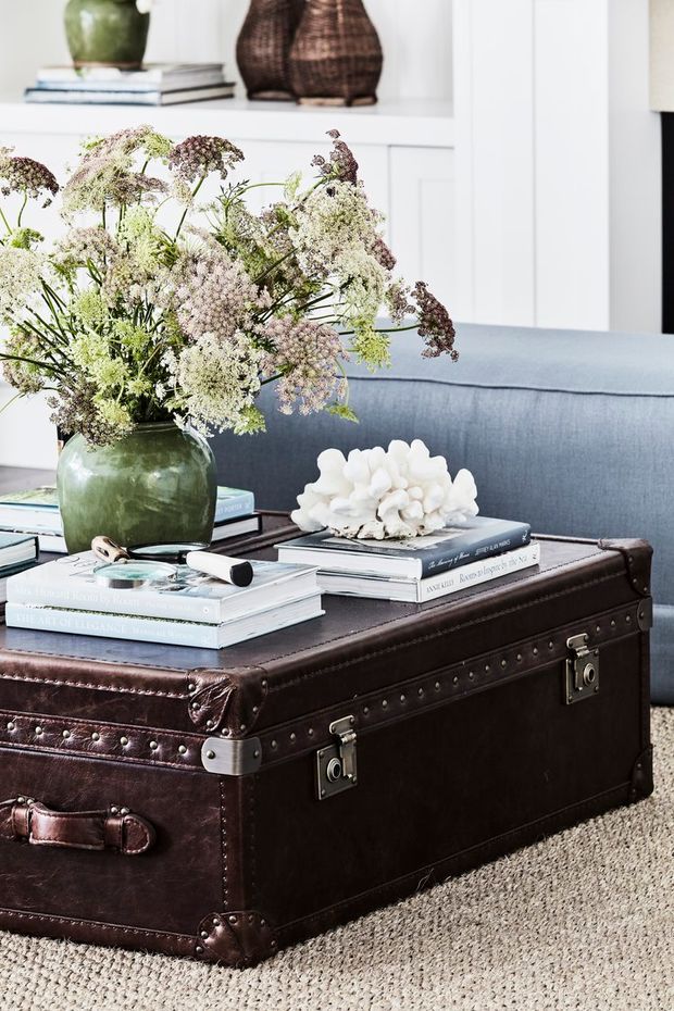 A styled living room vignette featuring a large vintage dark brown leather trunk used as a coffee table, stacked books, decorative objects, a green ceramic vase filled with dried flowers, and a blue upholstered sofa. White built-in shelving with books and woven vessels forms the background, while a neutral textured carpet anchors the room.