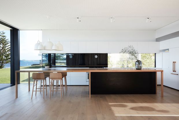 A bright contemporary kitchen with a large timber-framed island and dark front panel, white flat-panel cabinetry, a dark glossy splashback, timber flooring, and three bar stools. Full-height glazing provides views toward a coastal landscape, while pendant lights and restrained styling create an airy, refined interior.