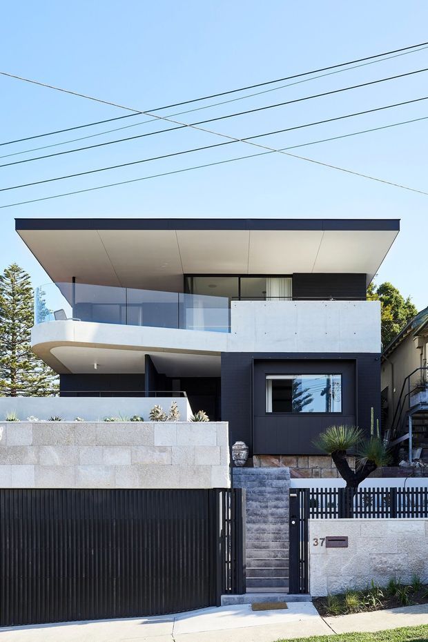 Street-facing photograph of a contemporary multi-level residence with a broad flat roof, projecting upper floor, curved balcony edge, clear glass balustrades, white rendered and dark charcoal facade sections, and a front boundary wall with integrated gate and garage door. Stone retaining walls, landscaping, neighbouring homes, and overhead utility wires are visible.
