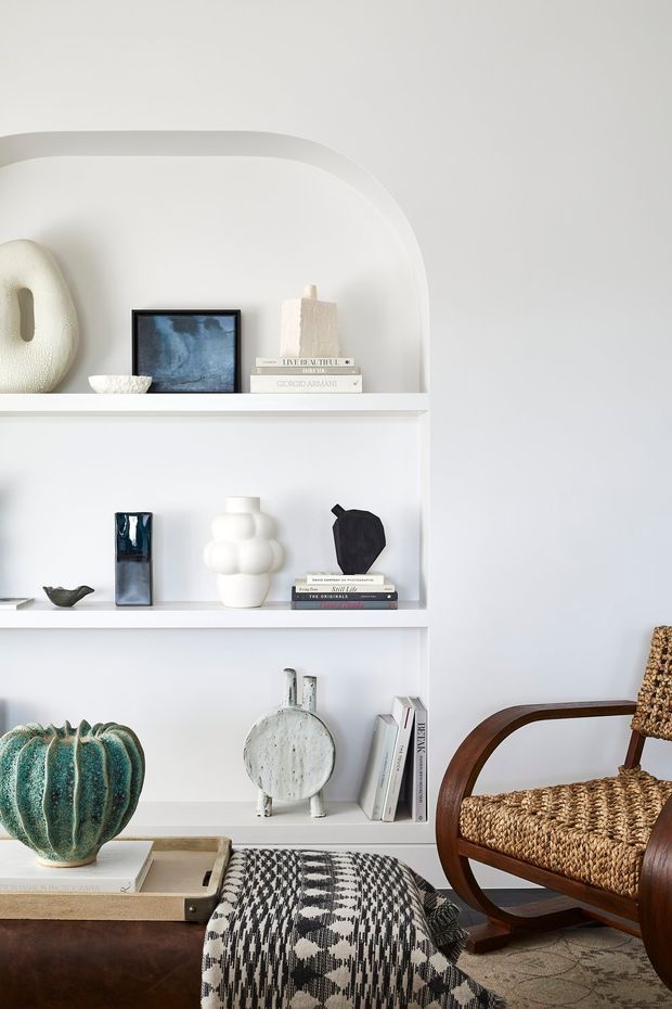 A styled interior vignette featuring white built-in arched shelving filled with books, ceramics, sculptural objects and framed artwork. A woven-seat wooden armchair sits beside a leather ottoman or bench draped with a black-and-white patterned textile. The room has a restrained white and neutral palette with sculptural decorative accents and a refined, curated feel.
