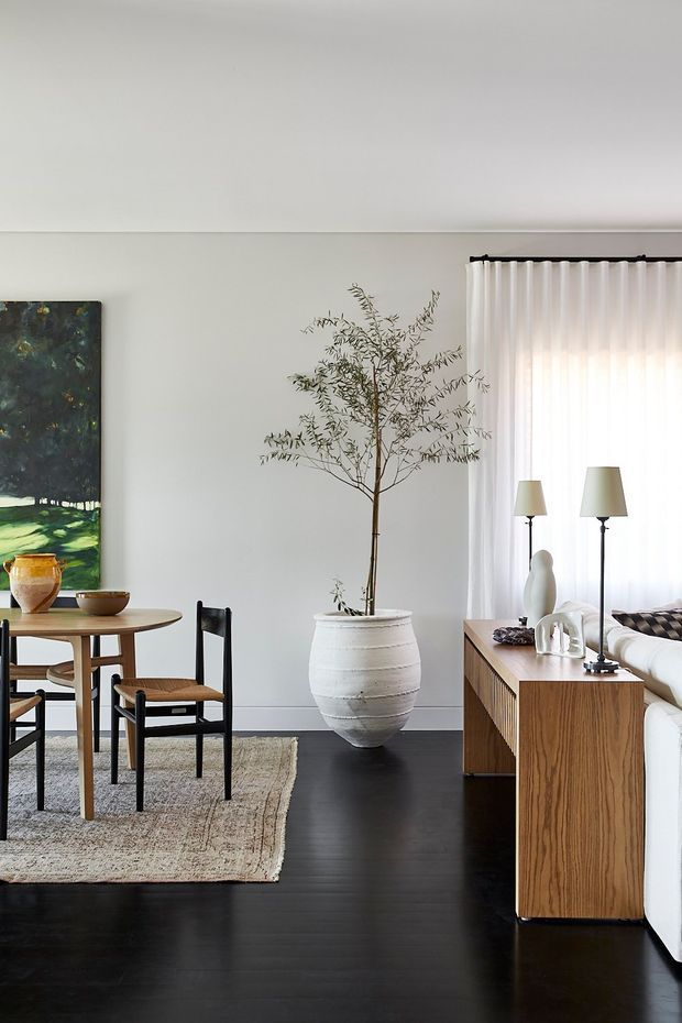 A refined open-plan dining and living area with a round timber dining table, black-framed upholstered chairs, a textured neutral rug, dark timber flooring, and a large potted indoor tree. A timber sideboard with decorative objects and twin table lamps sits beside a partially visible white sofa. White walls, sheer floor-to-ceiling curtains, and contemporary artwork create a calm, restrained interior.