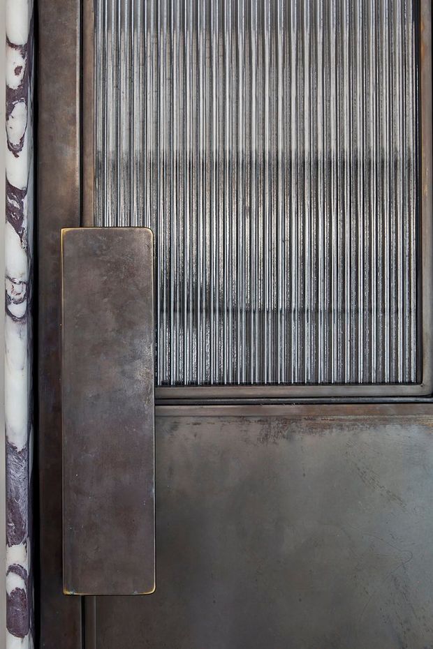Close-up detail of an industrial-style metal door or architectural panel. The image shows a weathered dark metal surface, a large rectangular bronze-brown metal pull handle, a narrow vertically ribbed translucent or reflective panel, and a contrasting marbled edge strip. The composition emphasizes tactile materiality, patina, and linear texture.