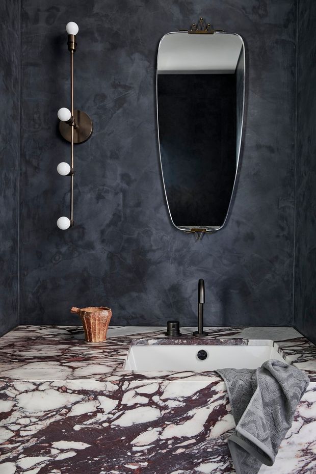 A dramatic, compact bathroom vanity featuring a white undermount basin set into a richly veined burgundy-and-white marble countertop and splashback. The walls are finished in dark charcoal textured plaster, complemented by a slim black tap, dark basin hardware, a vintage-inspired sculptural mirror, and a vertical globe-bulb wall light. A folded grey towel and small copper-toned vessel add practical and decorative details.