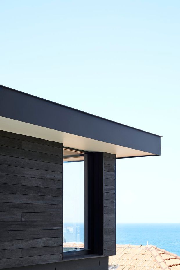 Close-up exterior view of a contemporary coastal building corner, featuring dark horizontal cladding, a large recessed window with dark framing, and a broad flat roof overhang. Blue ocean and neighbouring terracotta roof tiles are visible beyond the facade.