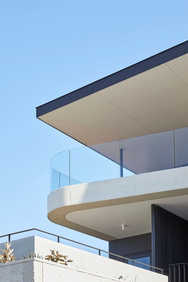 Close-up upward view of a contemporary multi-level building exterior with a dramatic cantilevered flat roof, broad projecting eaves, curved concrete balcony, frameless glass balustrade, dark vertical screening, and pale rendered/concrete surfaces against a clear blue sky.