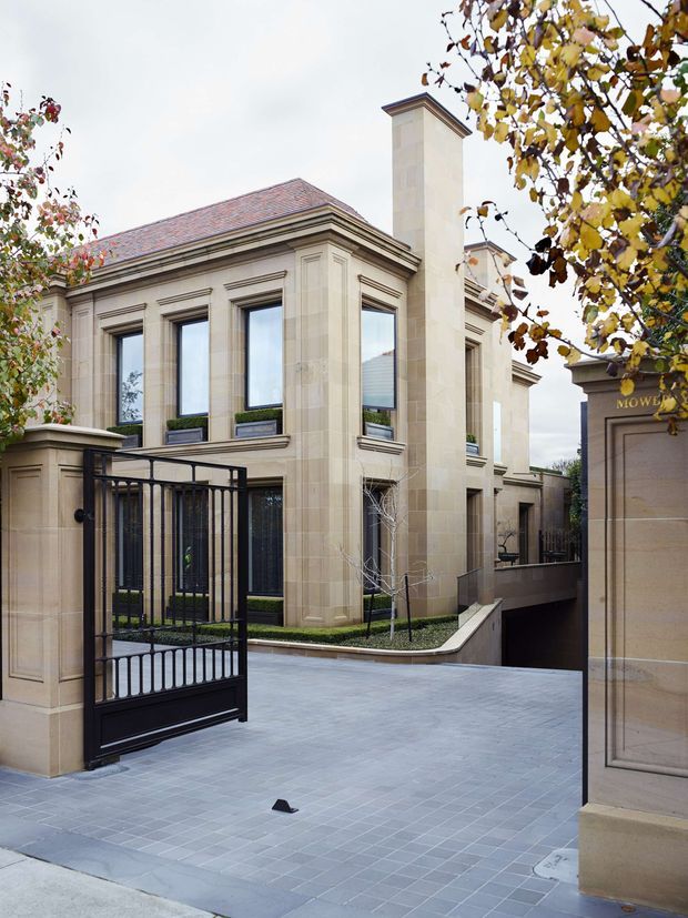 Exterior view of a formal, classically detailed stone-clad residence behind a black metal security gate. The building has tall recessed windows with strong moulded surrounds, a hipped terracotta-tile roof, prominent chimney, landscaped courtyard planting, and a broad grey paved driveway or entry court. The composition feels refined and grand, with mature deciduous trees framing the facade.