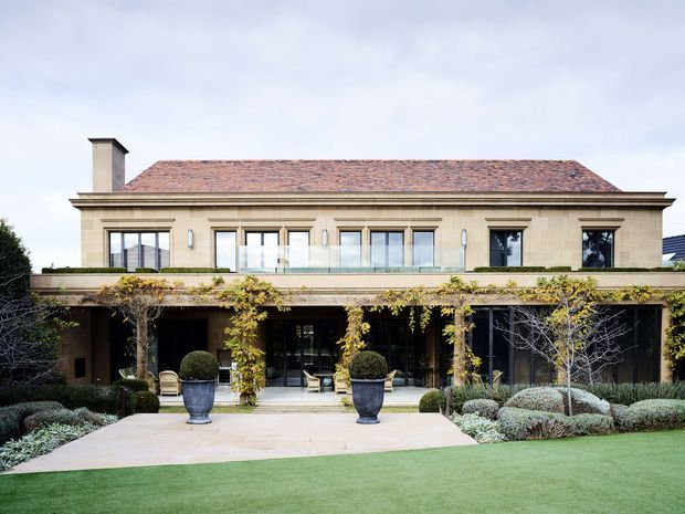 A formal two-storey residential house viewed from the landscaped front garden. The symmetrical rendered facade has classical detailing, tall multi-pane windows and doors, a broad upper-level balcony or terrace, and a low-pitched terracotta tiled roof with chimney. A deep ground-floor veranda is supported by slender columns and partly covered with climbing vines. The foreground features clipped hedges, urn planters, pale stone paving and a manicured lawn.
