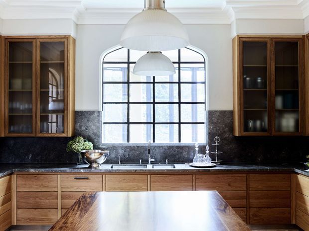 A bright, refined kitchen with symmetrical built-in timber cabinetry, dark stone splashback and benchtops, a large black-framed gridded window, and a central timber island. White walls with decorative ceiling cornices and oversized dome pendant lights create a classic architectural backdrop with warm contemporary finishes.