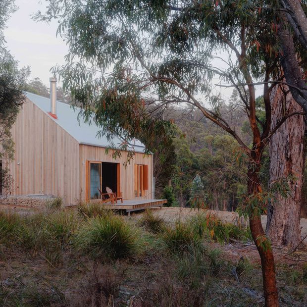 A compact single-storey timber-clad cabin set among native bushland, with a simple pitched metal roof, chimney, and a small raised timber deck accessed through glazed sliding doors. Mature eucalyptus trees and natural rocky terrain surround the dwelling, creating a secluded woodland setting.