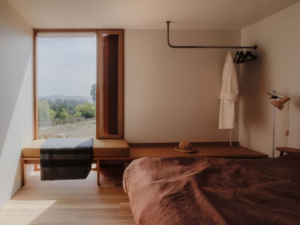 Warm, minimalist bedroom with a low timber platform bed, rumpled brown bedspread, built-in timber window seat, and a compact wall-mounted clothing rail. The room has smooth warm off-white walls, light timber flooring, and a deep timber-framed window overlooking a leafy landscape. A simple floor lamp and a few understated furnishings reinforce the calm, pared-back aesthetic.