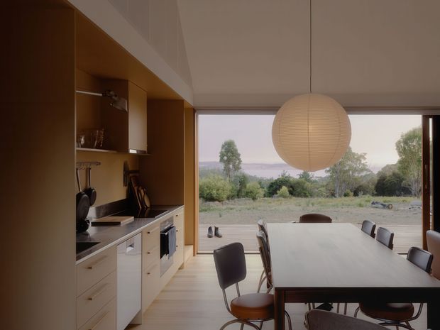 Warm, contemporary open-plan kitchen and dining area with light timber flooring, white built-in cabinetry, and a dark wood or stone kitchen worktop. Open shelving and integrated appliances line the left wall, while a large dining table with mixed dark and brown chairs sits beside expansive glazing and an outdoor view. A large spherical pendant light hangs above the table. The space has a restrained mid-century-influenced feel with strong indoor-outdoor connection.