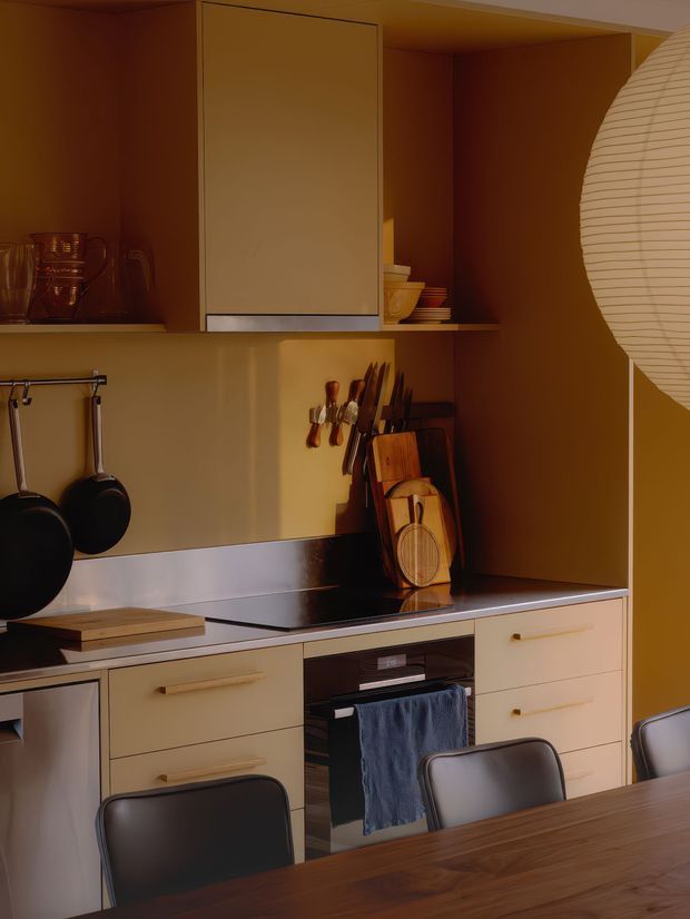 Warm, compact contemporary kitchen viewed from the dining area, featuring flat-panel tan cabinetry, integrated appliances, a dark cooktop and splashback, open shelving with crockery, hanging cookware, knife storage, timber chopping boards, and a large softly glowing paper pendant at the right. A dark timber dining table and upholstered chairs occupy the foreground.