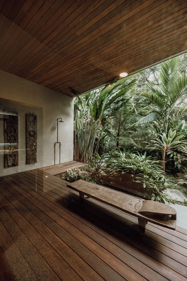A refined tropical indoor-outdoor bathing area opening onto dense lush planting. The space features a broad dark timber deck, matching slatted timber ceiling, a built-in stone or concrete bench, and a minimalist wall-mounted shower with a freestanding-style tap fitting. Decorative carved panels and a patterned screen are visible on the wall, while abundant palms and tropical foliage fill the courtyard beyond.