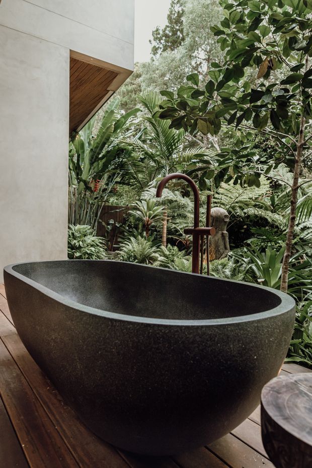 An open-air bathroom featuring a large freestanding dark stone bathtub on a timber deck, with an exposed timber-lined roof overhang, concrete-rendered wall, and lush tropical garden surrounding the bathing area. A sculptural dark metal bath tap and matching freestanding fixture sit behind the tub.