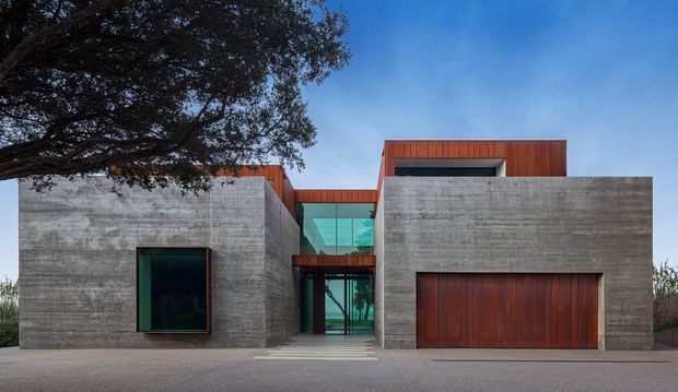 Front exterior view of a contemporary, rectilinear residence composed of exposed horizontal-board concrete volumes, warm red-brown timber-clad upper sections, a large timber garage door, and a central turquoise-tinted glazed entry link. A mature tree partially frames the facade.