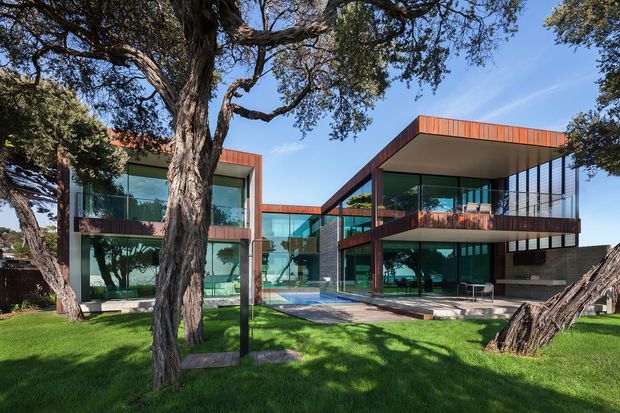 A contemporary multi-level house set among mature trees, with extensive floor-to-ceiling glazing, strong horizontal volumes, cantilevered upper levels, and warm rust-coloured metal cladding. The residence opens onto a manicured lawn and appears to include a central outdoor pool or water feature visible through the glass.