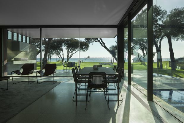 A sophisticated modern open-plan living and dining space with floor-to-ceiling glazing overlooking a landscaped lawn, trees, and a swimming pool. The room features a dark dining table with wire-frame chairs, sculptural lounge chairs, polished concrete flooring, and a minimalist built-in fireplace or feature wall. Strong indoor-outdoor connection and expansive views define the space.