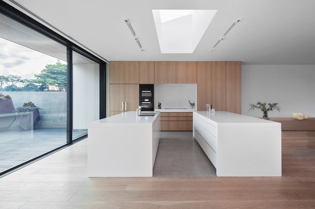 Bright contemporary kitchen with two large white waterfall islands, integrated white cabinetry, warm timber wall panelling and flooring, and a dark stone or composite benchtop at the rear. Full-height black-framed sliding glazing opens to an outdoor terrace, while a large skylight and linear ceiling lights provide natural and artificial illumination.