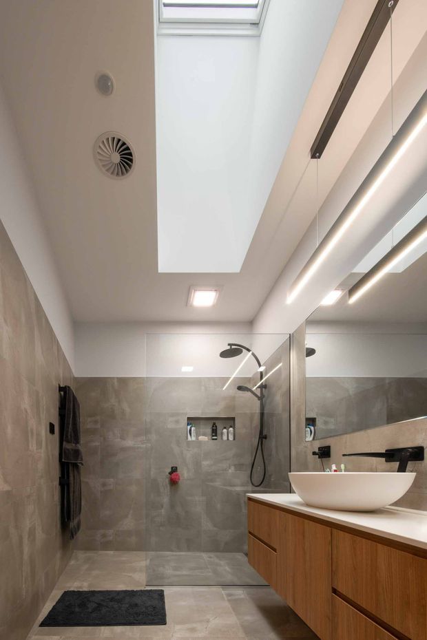 A contemporary bathroom with a walk-in glass shower, wall-mounted dark fixtures, a white vessel basin on a timber vanity, large grey stone-look wall and floor tiles, mirrored cabinetry or wall surfaces, linear LED lighting, recessed ceiling lights, and a skylight.
