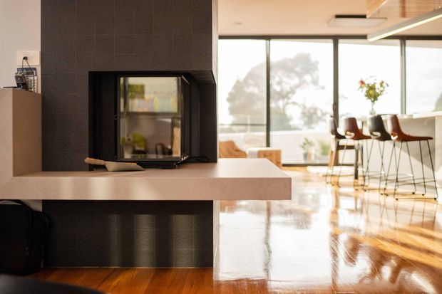 Warm contemporary open-plan interior centred on a freestanding fireplace clad in dark textured tile, with a pale stone projecting hearth or bench. Beyond it is a bright kitchen and dining area with floor-to-ceiling glazing, bar seating, timber cabinetry, and polished timber flooring reflecting the daylight.