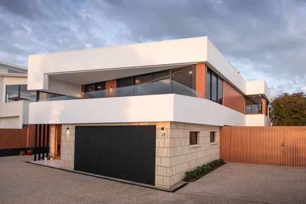 Exterior view of a contemporary multi-level residence with a clean flat-roofed form, white rendered upper volumes, warm timber-toned vertical cladding, a glass-balustraded balcony, and a dark integrated garage door. The lower facade uses light masonry blockwork, with a timber privacy fence and driveway in the foreground.