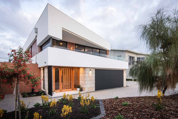 Exterior view of a contemporary two-storey residence with crisp white geometric volumes, a projecting upper level, recessed balcony, dark garage door, timber entry screen, and extensive glazing. The home is set within a landscaped suburban frontage with paved access and garden beds.
