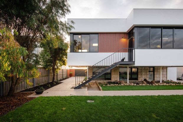 Exterior photograph of a modern two-storey residence with a crisp white rendered upper volume, extensive dark-framed glazing, a vertical timber-clad accent wall, and an exposed external steel stair leading to an elevated entry. The lower level combines pale masonry walls with open parking or undercroft space. A landscaped front lawn, concrete path, timber boundary fencing, and established trees surround the house.