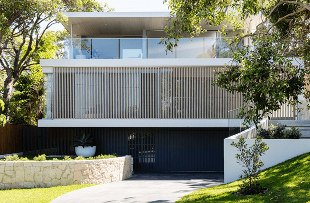 Front exterior view of a contemporary multi-level residence framed by mature trees and landscaped gardens. The facade combines white rendered walls, extensive upper-level glazing with a glass balustrade, a broad vertical slatted privacy screen, and a dark recessed garage or entry level. A pale stone retaining wall and curved driveway complete the foreground.